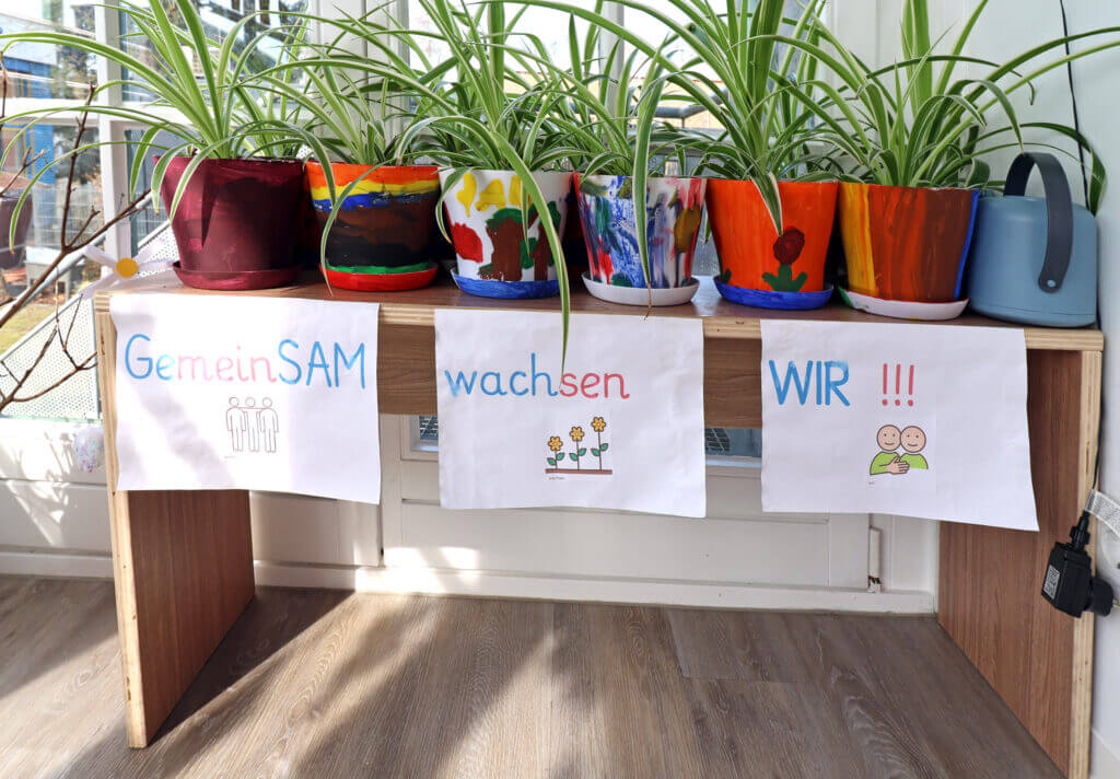 Sachunterricht – Pflanzen in bemalten Blumentöpfen, darunter ist auf Papier geschrieben „GemeinSAM wachsen wir!!!“